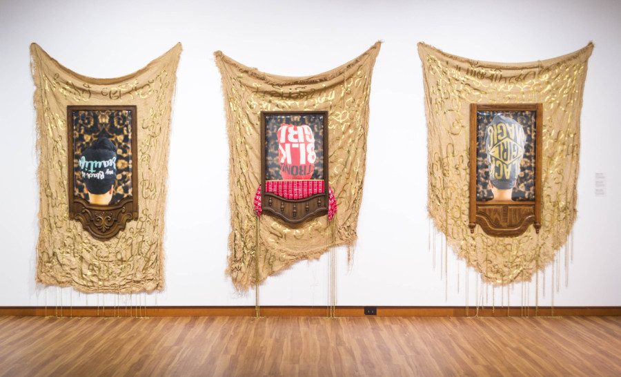 A Radiant Revolution III, II, and I by Stephanie J. Woods, 2018, Burlap dyed with sweet tea, woven brass chains, t-shirt, textile foil, polished furniture vinyl, dresser mirror frame, and upholstered taffeta print

Inspired by graphic t-shirts featuring phrases such as “My Black is Beautiful,” Strong Black Girl,” and “Black Girl Magic.” The t-shirts are captured in a series of photographs that relate these expressions of empowerment to the history of head wraps. There was a time in history when sumptuary laws banned black women from showing our hair. However, the head wrap then and now represents courage, ancestry, collective identity, and a uniform of rebellion that signifies the resistance to loss of self-definition. Each photograph is upholstered inside of an inverted dresser mirror frame and protected with polished furniture vinyl. Behind each photograph drapes hand-sewn burlap tapestries that are dyed with sweet tea giving the pieces a copper color. The burlap tapestries are also embellished with gold scrambled text.